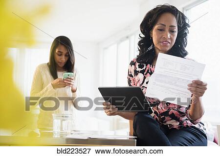 Stock Photo - Mother paying bills with digital tablet while daughter uses cell phone. Fotosearch