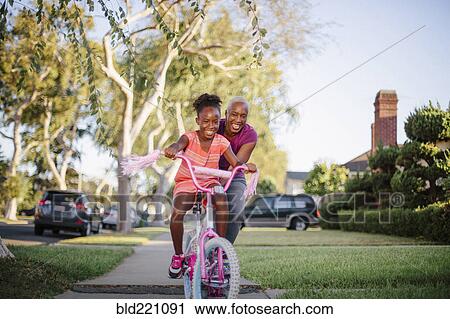 Stock Image - Mother pushing daughter on bicycle. Fotosearch