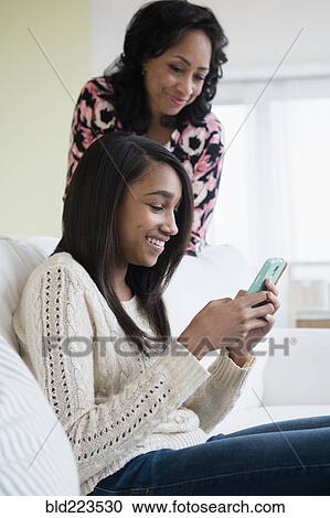 Stock Image - Mother watching daughter use cell phone on sofa. Fotosearch