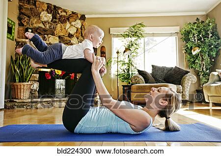 Stock Image - Mother working out on exercise mat balancing baby on legs. Fotosearch