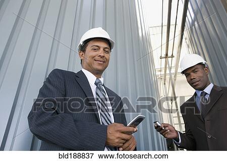 Multi-ethnic businessmen in hard-hats holding cell phones View Large Photo Image Stock Photo - Multi-ethnic businessmen in hard-hats holding cell phones. Fotosearch