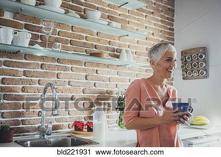Stock Image - Older Caucasian woman having coffee in kitchen. Fotosearch