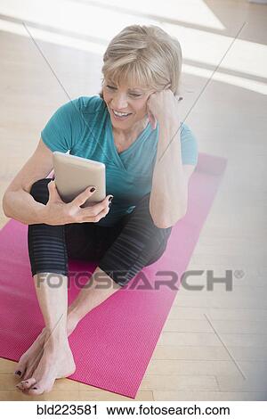 Stock Image - Older Caucasian woman on exercise mat using digital tablet. Fotosearch