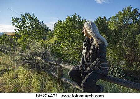 Stock Image - Older Caucasian woman sitting on wooden fence. Fotosearch