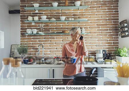 Stock Image - Older Caucasian woman talking on cell phone in kitchen. Fotosearch
