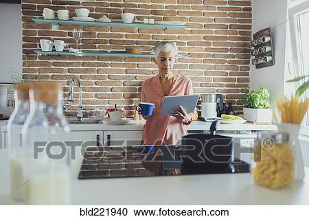 Stock Image - Older Caucasian woman using digital tablet in kitchen. Fotosearch