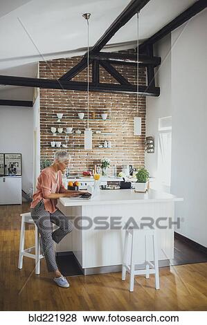 Stock Photo - Older Caucasian woman using laptop in kitchen. Fotosearch