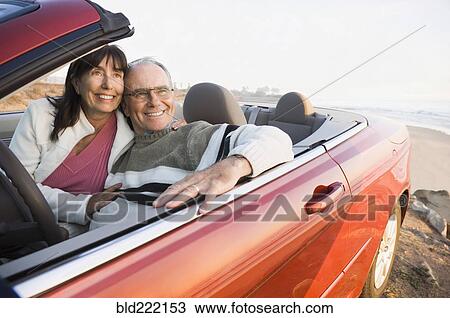 Stock Image - Older couple driving convertible on beach. Fotosearch