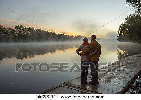 Stock Image - Older couple hugging at foggy river at sunrise. Fotosearch