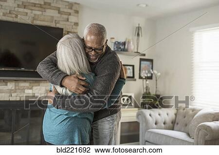 Stock Image - Older couple hugging in living room. Fotosearch