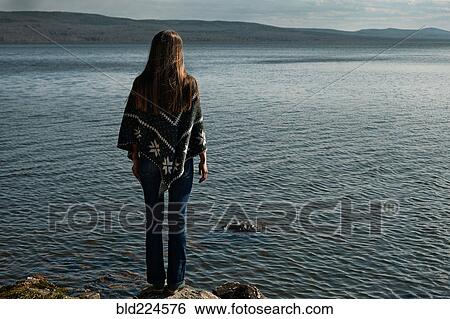 Stock Photograph - Pensive Caucasian girl standing on rock at lake. Fotosearch