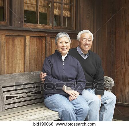 Stock Photograph - Senior Asian couple smiling and sitting on bench outdoors. Fotosearch