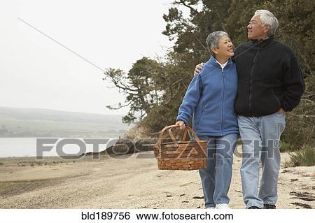 Stock Photograph - Senior Asian couple walking on beach with picnic basket. Fotosearch