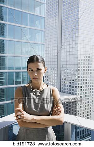 Stock Photo - Serious Hispanic woman standing on urban balcony with arms crossed. Fotosearch