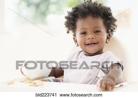 Stock Image - Smiling Black baby girl eating cereal from bowl in high chair. Fotosearch