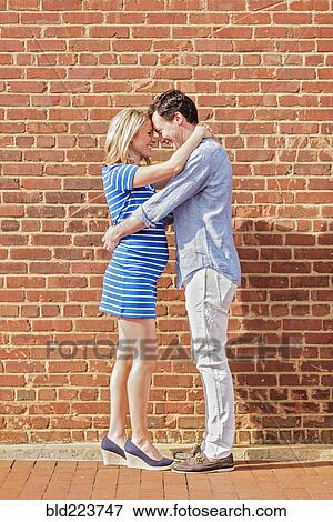 Stock Photo - Smiling Caucasian man and expectant mother hugging near red brick wall. Fotosearch