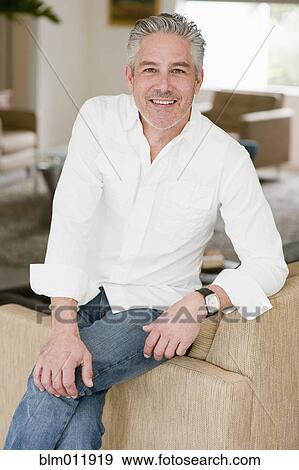 Stock Photo - Smiling man sitting on sofa in living room. Fotosearch