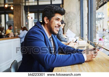 Stock Image - Smiling Middle Eastern man using cell phone at cafe window. Fotosearch