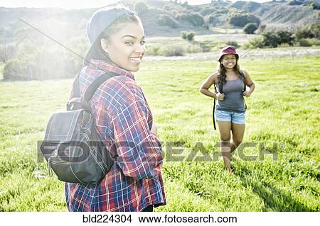 Picture - Smiling Mixed Race sisters backpacking in field near mountain. Fotosearch