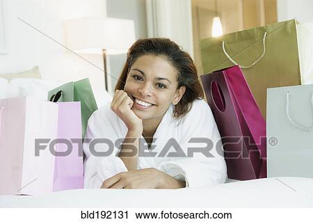 Stock Image - Smiling mixed race woman laying on bed with shopping bags. Fotosearch