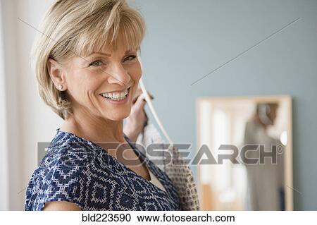 Stock Image - Smiling older Caucasian woman holding clothing near mirror. Fotosearch