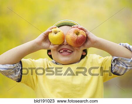 Stock Image - Toothless Hispanic boy covering eyes with apples. Fotosearch
