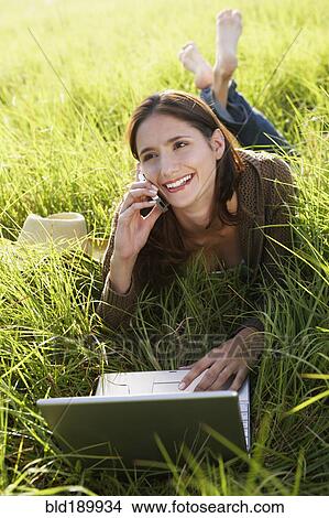 Picture - Woman laying in meadow using laptop and cell phone. Fotosearch
