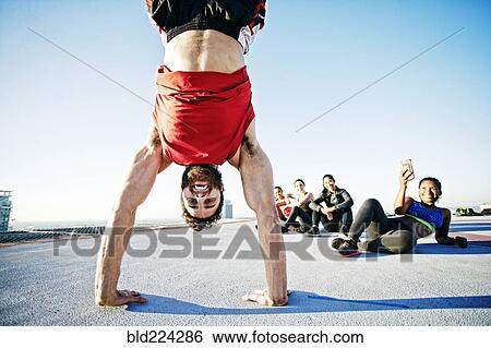 Stock Photograph - Woman photographing man doing handstand on urban rooftop. Fotosearch