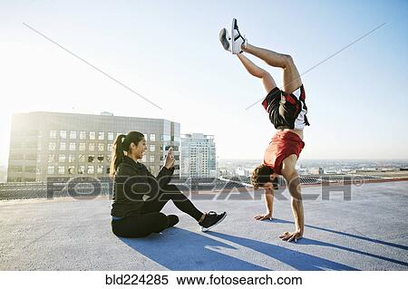 Stock Photography - Woman photographing man doing handstand on urban rooftop. Fotosearch