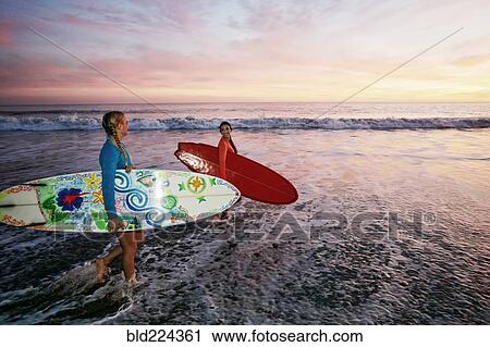 Stock Image - Women carrying surfboards in ocean waves at beach. Fotosearch