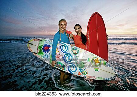 Stock Image - Women standing in ocean waves holding surfboards at beach. Fotosearch