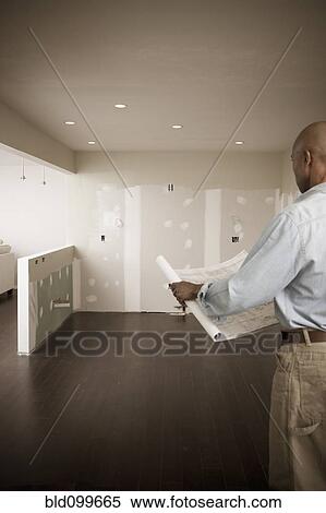 Stock Photography - African American man standing in unfinished room with blueprints. Fotosearch