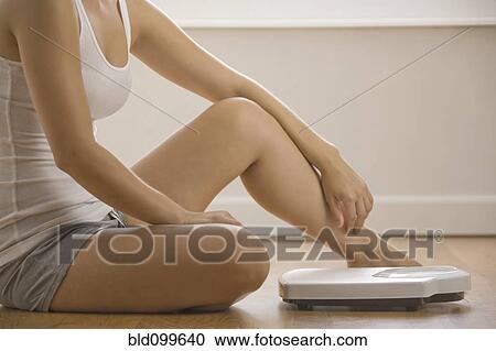 Stock Image - Caucasian woman sitting on floor near scale. Fotosearch