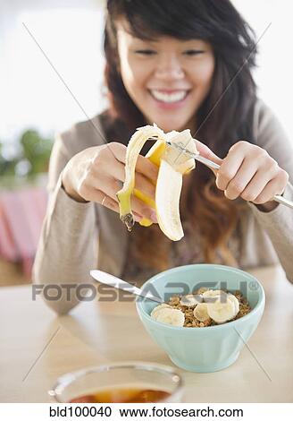 Stock Image - Pacific Islander woman putting bananas on cereal. Fotosearch
