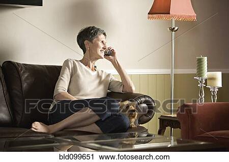 Stock Image - Senior woman sitting on sofa talking on telephone. Fotosearch