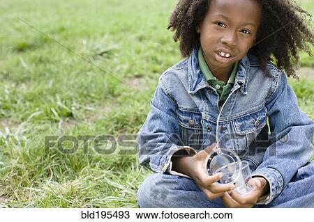 Stock Image - African American boy sitting in grass and holding jar. Fotosearch