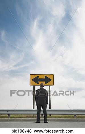 Stock Photo - African American businessman looking at conflicting road sign. Fotosearch