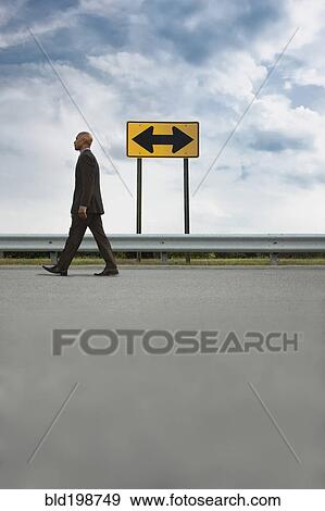 Stock Photo - African American businessman walking on road near road sign. Fotosearch