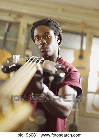 Stock Image - African American craftsman looking at guitar in workshop. Fotosearch