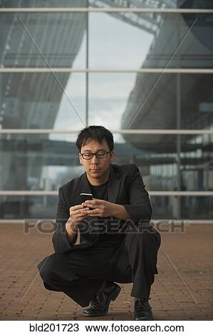 Stock Image - Asian businessman squatting and text messaging on cell phone. Fotosearch