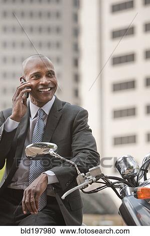 Stock Image - Black businessman sitting on scooter talking on cell phone. Fotosearch