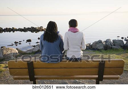 Stock Image - Black couple sitting on bench near shore. Fotosearch