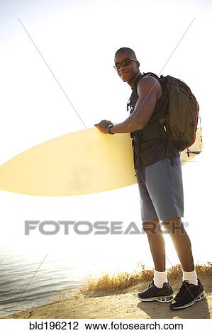 Stock Image - Black man carrying surfboard near ocean. Fotosearch