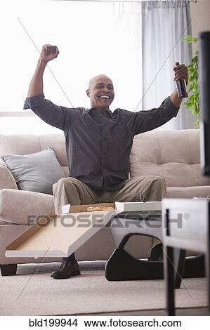 Picture - Black man cheering at television and eating pizza. Fotosearch