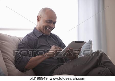 Stock Photo - Black man sitting on sofa using digital tablet. Fotosearch