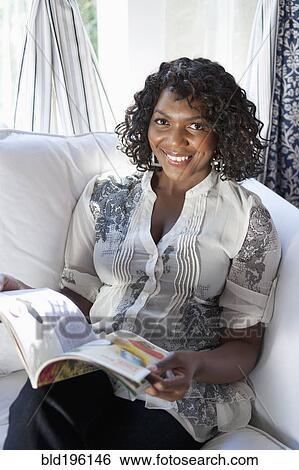 Stock Photograph - Black woman reading magazine on sofa. Fotosearch