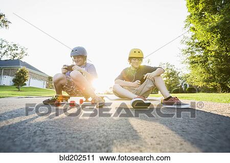 Stock Image - Caucasian boys sitting on skateboards and using cell phones. Fotosearch