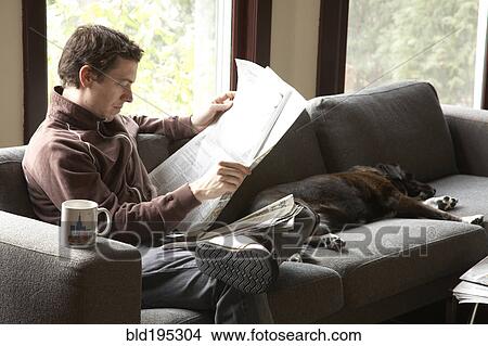 Picture - Caucasian man reading newspaper on sofa with dog. Fotosearch