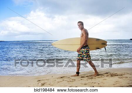 Picture - Caucasian man walking on beach carrying surfboard. Fotosearch