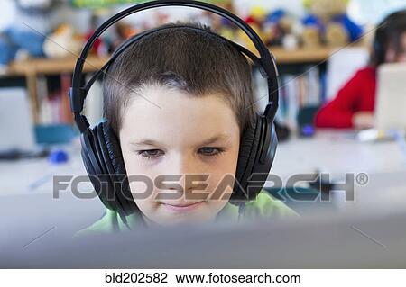 Stock Image - Caucasian student in headphones using laptop in classroom. Fotosearch